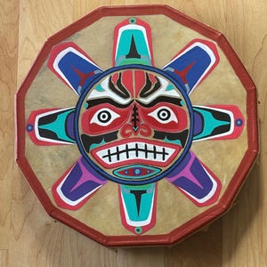 May include: A round drum with a red octagonal frame and a painted sun design. The sun has a red face with white eyes and teeth, surrounded by blue, green, and purple rays. The drum is tan in color.