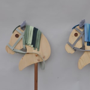 Op de afbeelding: Twee houten stokpaarden met lichtbruine lichamen, grijze teugels en kleurrijke franje manen. Eén heeft groene en marineblauwe franje, de andere blauw. De paarden staan op houten palen.