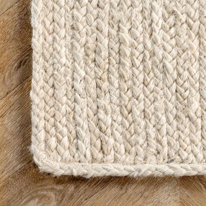 Puede incluir: Primer plano de una alfombra tejida natural con una textura trenzada. La alfombra es de color beige claro y tiene un aspecto grueso y duradero. El borde de la alfombra está cuidadosamente terminado con un borde trenzado. La alfombra está colocada sobre un suelo de madera.