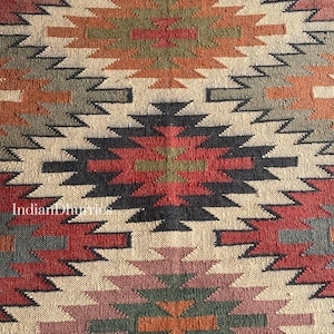 May include: Close-up of a woven rug with a geometric pattern. The rug features a repeating design of diamond and zigzag shapes in shades of red, orange, green, black, and beige. The text "Indian Dhurries" is visible in the lower left corner.