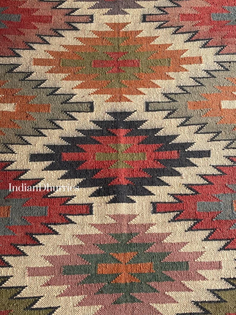 May include: Close-up of a woven rug with a geometric pattern. The rug features a repeating design of diamond and zigzag shapes in shades of red, orange, green, black, and beige. The text "Indian Dhurries" is visible in the lower left corner.