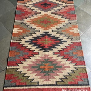 May include: A rectangular rug with a geometric pattern in shades of red, beige, gray, and green. The rug has a fringed edge and the text "IndianDhurries" is visible. The rug is made of a woven material.