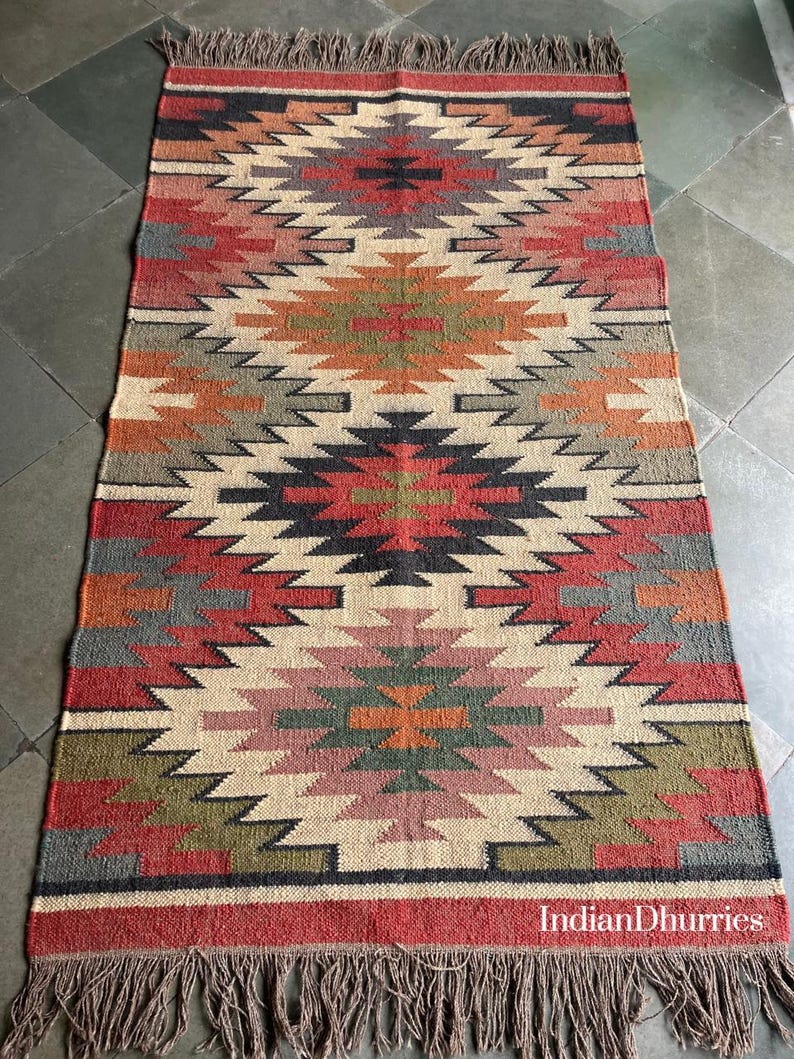 May include: A rectangular rug with a geometric pattern in shades of red, beige, gray, and green. The rug has a fringed edge and the text "IndianDhurries" is visible. The rug is made of a woven material.