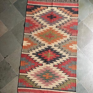 May include: Rectangular rug with a geometric pattern in shades of red, orange, beige, gray, and green. The rug has a fringed edge and a symmetrical design. The rug is suitable for a living room or bedroom.