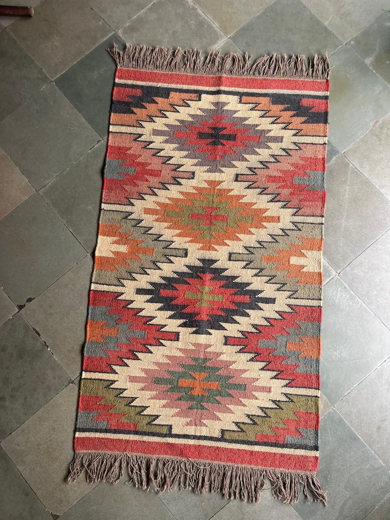 May include: Rectangular rug with a geometric pattern in shades of red, orange, beige, gray, and green. The rug has a fringed edge and a symmetrical design. The rug is suitable for a living room or bedroom.