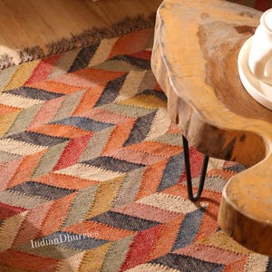 May include: A colorful area rug with a geometric chevron pattern in shades of orange, red, blue, and beige. The rug has a fringed edge and the text "Indian Dhurries" is visible. A wooden table with a live edge is in the background.