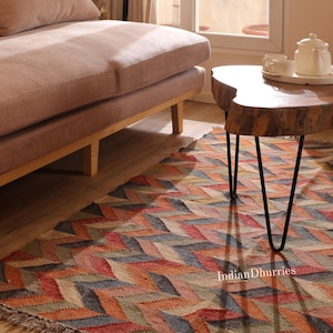 Op de afbeelding: Een kleurrijk vloerkleed met een chevronpatroon in tinten rood, oranje, blauw en beige. Het vloerkleed ligt op een houten vloer, met een bank en een houten salontafel met zwarte haarspeldpoten op de achtergrond. De tekst "Indian Dhurries" is zichtbaar.