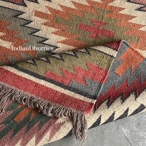 May include: A woven rug with a geometric pattern in shades of beige, red, orange, green, and black. The rug has a fringed edge and the text "Indian Dhurries" is visible.