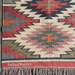 May include: Woven jute rug with a geometric pattern in red, black, cream, green, and orange. The rug has a fringed edge and the text "Indian Dhurries" is visible. This rug is suitable for home decor.