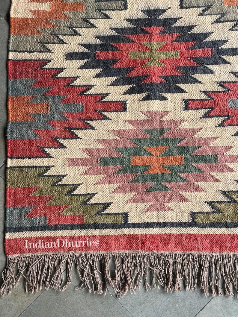 May include: Woven jute rug with a geometric pattern in red, black, cream, green, and orange. The rug has a fringed edge and the text "Indian Dhurries" is visible. This rug is suitable for home decor.