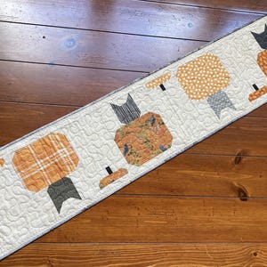 May include: A rectangular quilted table runner featuring a cream-colored background with a meandering stitch pattern. The runner displays four appliqued pumpkin shapes in various orange and brown fabric patterns, each topped with a dark gray stem.