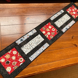 May include: A black and white table runner with a repeating pattern of coffee cups and musical notes. The runner has four red squares with a pattern of coffee cups on a red background.