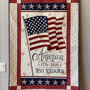 Puede incluir: Un edredón patriótico con la bandera estadounidense, con rayas rojas y blancas y un campo de estrellas azul. El edredón tiene un fondo blanco, bordes rojos y azules y el texto "America 1776-2026 250 YEARS".