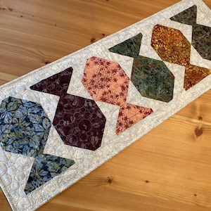 May include: A quilted table runner featuring a repeating fish design. The fish are made from various patterned fabrics in shades of blue, burgundy, orange, and green. The runner is bordered with a white fabric and quilted with a circular pattern.