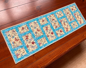 Puede incluir: Un camino de mesa acolchado azul y crema con un patrón de mariposa. El camino de mesa tiene un borde azul y está hecho de tela.