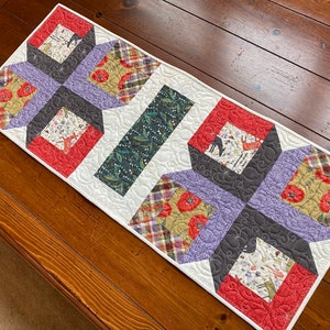 May include: A quilted table runner with a geometric pattern in shades of red, green, purple, and white. The runner features a central cross shape with smaller squares surrounding it.