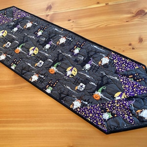 Può includere: Un runner da tavolo di Halloween nero e viola con un design trapuntato. Il runner presenta un motivo ripetitivo di streghe, zucche e case. Il runner ha un bordo nero.