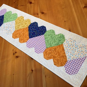 Op de afbeelding: Een witte tafelrunner met een rij kleurrijke patchwork harten. De harten zijn gemaakt van verschillende stoffen met bloemen-, ruit- en geometrische patronen. De runner is gequilt met wit garen.