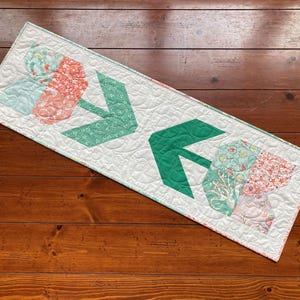 Puede incluir: Un camino de mesa blanco con un patrón floral verde y rosa. El camino de mesa presenta dos flechas verdes grandes que apuntan hacia la derecha.