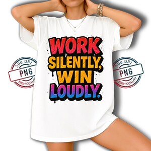 May include: White t-shirt with the text "WORK SILENTLY, WIN LOUDLY" in a colorful, graffiti-style design. The text is in red, yellow, and blue, with black accents. The shirt has short sleeves and a relaxed fit.