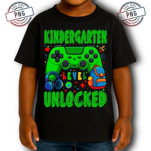 May include: Black t-shirt with a green graphic that says "Kindergarten Level Unlocked." The graphic includes a green video game controller, headphones, a backpack, and other colorful elements. The shirt is designed for children.