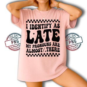 May include: A light pink t-shirt with the text "I IDENTIFY AS LATE MY PRONOUNS ARE ALMOST/.THERE" in black, with a checkerboard pattern above and below the text. The shirt has short sleeves.