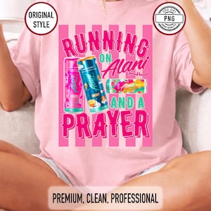 May include: Pale pink t-shirt featuring the slogan "RUNNING ON Alani AND A PRAYER" in pink and white. The design incorporates images of Alani Nu energy drink cans. The top has a round neck and short sleeves.