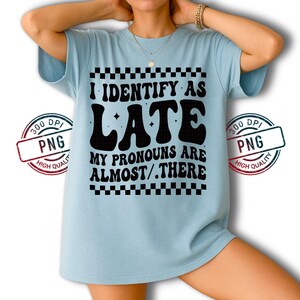 May include: Light blue t-shirt with the text "I IDENTIFY AS LATE MY PRONOUNS ARE ALMOST/.THERE" in black. The text is framed by a black and white checkered pattern. The shirt is a casual wear item.