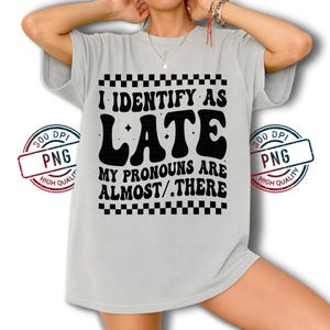 May include: A light gray t-shirt with the text "I IDENTIFY AS LATE MY PRONOUNS ARE ALMOST/.THERE" in black. The text is in a retro font with a checkered pattern above and below the text. The shirt has short sleeves.