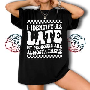 May include: Black t-shirt with the text "I IDENTIFY AS LATE. MY PRONOUNS ARE ALMOST/.THERE" in white. The text is in a retro, wavy font, with a checkered pattern above and below the text.