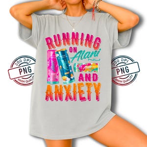 May include: A light gray t-shirt with the text "RUNNING ON Alani AND ANXIETY" in a colorful, retro font. The shirt features three cans of Alani Nu energy drinks in pink, blue, and a pink and green design.