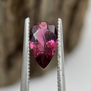Puede incluir: Una piedra preciosa facetada en forma de pera, de color magenta y rosa intenso, sostenida por unas pinzas de metal plateado. La piedra presenta múltiples facetas triangulares y en forma de cometa que reflejan la luz con transparencia.