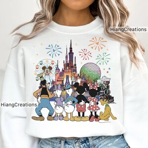 以下が含まれることがあります： ミッキーマウスとミニーマウスを含むディズニーのキャラクターとランドマークのカラフルなグラフィックが特徴の白いスウェットシャツ。デザインにはお城、花火、電車が含まれています。「HiangCreations」という文字が側面にあります。