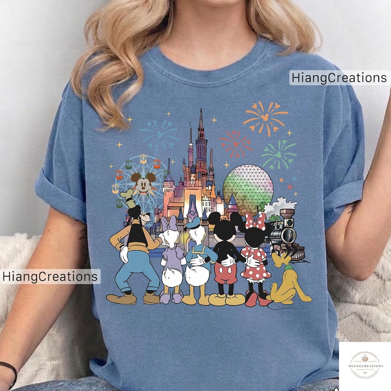 以下が含まれることがあります： 青いTシャツ。ディズニーのキャラクター、お城、花火、観覧車のカラフルなグラフィックが特徴です。「HiangCreations」の文字がプリントされています。