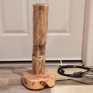 Puede incluir: Lámpara de madera artesanal con un acabado marrón claro natural. La lámpara presenta un diseño cilíndrico con una sección tallada y una base redonda. El cable de alimentación es negro.