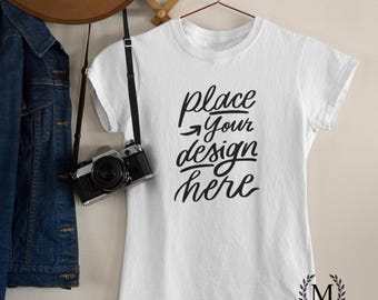 May include: White t-shirt with the text "place your design here" in black script. A vintage camera hangs from a strap next to the shirt. A denim jacket and a brown hat are also visible.