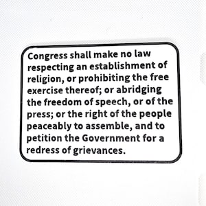 May include: A white background with a black and white rectangular sticker. The sticker contains text from the First Amendment of the U.S. Constitution, guaranteeing freedoms of religion, speech, press, assembly, and petition.