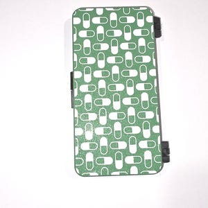 May include: A rectangular, hinged case with a green background and a repeating pattern of white pill shapes. The case has black hinges and a black clasp. The design is a modern, graphic style.