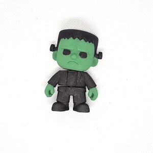 May include: A 3D-printed figurine of Frankenstein's monster. The figure has a green face, black hair, and a black suit. The figure is standing on a white surface.