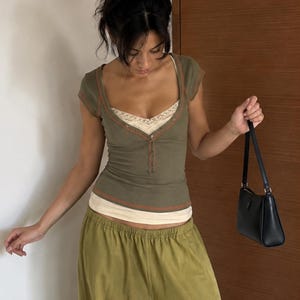 Peut inclure: Un haut henley à manches courtes vert olive avec une encolure dégagée, superposé sur un caraco crème bordé de dentelle. La tenue comprend un pantalon vert olive ample à taille élastique et un petit sac à main noir porté à l'épaule.