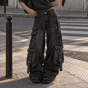 Puede incluir: Una camiseta gris carbón de hombros descubiertos con un gran gráfico de estrella blanca en el pecho, combinada con pantalones cargo negros holgados y desgastados. Los pantalones incluyen múltiples bolsillos laterales y hebillas.