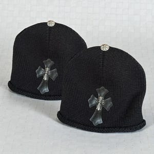 Pode incluir: Duas toucas pretas de malha com uma cruz prateada e um botão prateado no topo. A cruz é feita de prata e couro preto. As toucas são exibidas em uma superfície branca.