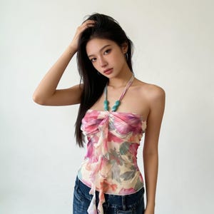 Könnte beinhalten: Ein ärmelloses Neckholder-Top aus transparentem, leichtem Stoff mit einem lebendigen Blumenmuster in Rosa-, Lila- und Grüntönen. Der Ausschnitt ist mit zwei großen, glatten, blaugrünen Perlen und einem kaskadierenden Rüschendetail vorne verziert.