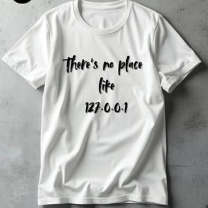 Op de afbeelding: Wit T-shirt met de tekst "There's no place like life 127.0.0.1" in zwart script. Het shirt heeft een ronde hals en korte mouwen. Een klein cirkelvormig logo staat in de linkerbovenhoek.