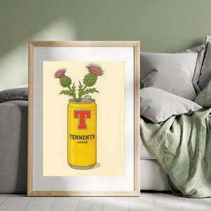 May include: A framed print featuring a yellow Tennent's Lager can with a red "T" logo, holding purple thistle flowers. The print has a cream-colored background and is displayed in a wooden frame. The image is set against a green wall.