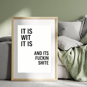 May include: A framed print with the text "IT IS WIT IT IS AND ITS FUCKIN SHITE" in black, centered on a white background. The frame is a light wood colour. The print is displayed on a wall near a bed with pillows and a green blanket.