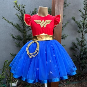 Könnte beinhalten: Ein Wonder Woman-Kostüm für Kinder. Das rote Oberteil hat goldene Verzierungen und ein goldenes Emblem. Der blaue Rock hat Sternakzente und ein goldenes Seil. Das Kostüm wird auf einer Schaufensterpuppe präsentiert.