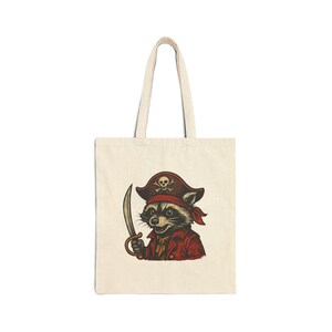 May include: Cream-colored tote bag featuring a cartoon raccoon dressed as a pirate. The raccoon wears a red pirate hat with a skull and crossbones, a red bandana, and a red coat. It holds a curved sword.
