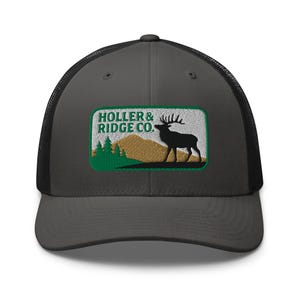 Puede incluir: Gorra de camionero gris carbón con la parte trasera de malla negra. La parte delantera presenta un parche rectangular con el texto "HOLLER & RIDGE CO." encima de una silueta de un alce, árboles y montañas en verde, dorado y negro.
