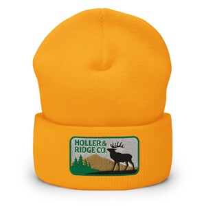 Pode incluir: Touca mostarda com aba dobrada. A frente apresenta um patch retangular com o texto "HOLLER & RIDGE CO." acima de uma silhueta de veado e montanhas em verde, marrom e preto.